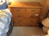 Full front view of the country style 4 drawer wooden dresser next to a bed, showing the natural wood finish and brass handles.