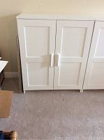 Front view of white IKEA Brimnes cupboard showing two panel doors with vertical handles on carpeted floor.