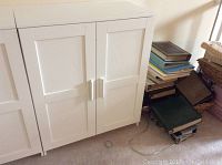 Front view of white IKEA Brimnes cupboard with two doors, placed next to a stack of books and basket.