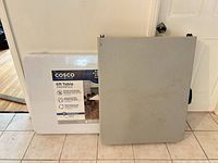 Cosco 6-ft folding table with label beside grey folding table, both folded