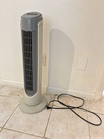 Full view of tower fan standing on tile floor showing front grille and power cord