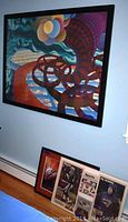 Photo showing large colorful abstract print with curved, patterned shapes hung on wall above two smaller framed pieces, one of which contains newspaper clippings.