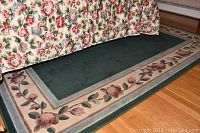 Close-up photo showing floral border and dark green center of rug under bed with floral linens.