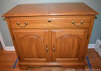 Front view of oak mobile server cabinet showing drawer, two cabinet doors, brass hardware, and carved base.