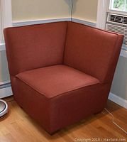 Rust-colored corner chair photographed from front-left angle in a corner next to a window with air conditioner.