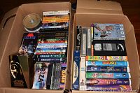 Box containing assorted DVDs and VHS tapes with some blank DVD-RW discs visible on top, various movie titles and covers.