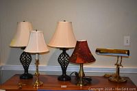 Photo showing five lamps: two tall dark lattice base table lamps with cream shades, brass candlestick lamp with cream shade, shorter lamp with reddish swirled shade, brass desk lamp