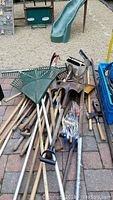 Overview of many garden tools stacked including various shovels, rakes, pruning tools, hand tiller, and other hand tools laid on ground outside on brick pavers.