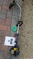 Full view of weed wacker showing motor, curved shaft, handle grip, circular cutting head with green cutting wire, protective guard, and manual booklet on the ground.