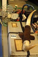 Shows vintage pale green metal tabletop fan, blue Dietz kerosene oil lamp behind it, decorative Mexican horn wall decor on top of beige laundry case, with visible texture.