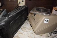 Closed plastic case marked Craftsman and beige plastic storage container for circular saw and blades near electrical outlet on marble surface.