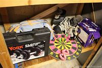 Photo of mixed sports and exercise equipment on a wooden shelf including the dumbbell set, dartboard, ice skates, and weights.