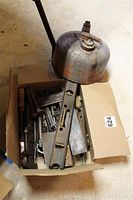 Box of assorted vintage metal hand tools including a rectangular wooden carpenter’s level, metal chisels, pliers, and other hand tools with a metal oil can placed on top.