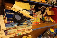 Stacked board games including Parchesi, Battleship, Monopoly, and The Game of Life with Yamaha digital percussion device and drumsticks on top. Various toys and puzzles visible around.