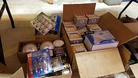 Overview photo of multiple open boxes containing various light bulbs and lighting accessories on a concrete floor under a table.