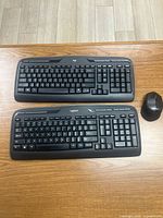 Two Logitech black wireless keyboards and one matching mouse on wooden surface