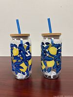 Front view of both lemon print glass tumblers with lids and straws
