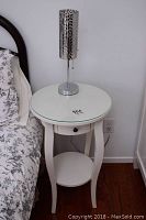 Tall white side table with round glass top, small drawer and lower shelf next to bed, lamp on top.
