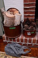 Two copper pots on brick hearth, larger pot filled with birch wood logs, smaller pot filled with pine cones, and a plastic bag on floor
