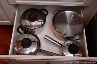 Four stainless steel pots with lids arranged in a drawer, showing various sizes and shapes with stainless steel and black knob lids.