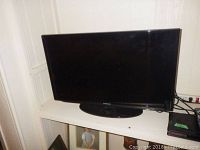 Front view of Samsung flat screen TV on a white shelf, black bezel and base stand visible.
