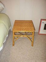 Front view of a square rattan side table with woven top and circular design on frame sides next to a bed and a framed picture on floor.