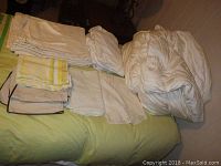 Multiple folded bedding linens including sheets and pillowcases in white, yellow stripe, beige with dark trim piled on a green bedspread.