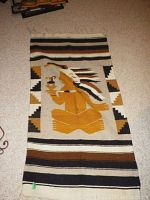 Woven rug with native American figure wearing a feathered headdress, holding a pipe, with geometric patterned borders in tan, brown, black, and white colors, laid flat on floor.