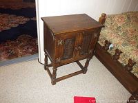 Full view of oak wood cabinet standing beside floral couch on carpeted floor. Shows front carved doors panel with brass hinges and knobs.