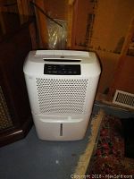 Front view of white Kenmore dehumidifier on floor in unfinished room, showing front grille and water tank window.