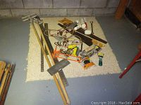 Photo of assorted hand tools including saw, silicone gun, plumbing tools and more, laid on a light-colored rug on concrete floor.