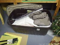 Bin of bedding and linens on floor, includes folded white linens, gray fabric with white circles, and a roll of curtain material.