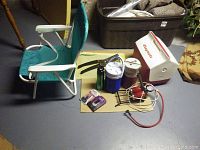 Photo of green/white folding camping chair, white/red Coleman Playmate cooler, assorted thermoses, shark deterrent device with cables and small camping items on floor.