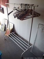 Full view of white metal clothing rack with assorted hangers hanging from the bar and shoe rack at bottom.