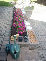 Overview of garden decor items including green elephant-shaped watering can, two black lanterns, stepping stones, and garden plaques arranged on stone garden bed edge beside a paved walkway with pink flowers.