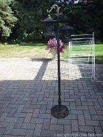 Full view of iron lantern post standing on paved surface outdoors, with two hanging lanterns and greenery background.