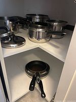 Group of stainless steel pots with lids and a pair of fry pans inside corner cabinet