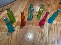 All seven colored glass bud vases on wood floor