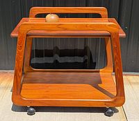 Front view of teak bar cart showing trapezoidal sides, lower shelf, casters
