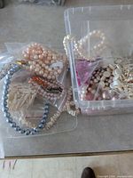 Overview of clear plastic container filled with assorted faux pearl strands, shell chip beads, loose beads and separate grey-blue bead strand