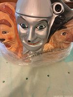 Front view of cookie jar showing Cowardly Lion, Tin Man, Scarecrow faces still wrapped in plastic
