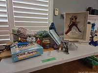 Overall lot on table showing board game, utensils, faux flowers, glass cake stand, figurine box