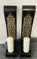 Front view of both black metal sconces with gold scroll pattern and included white pillar candles