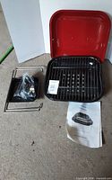 All grill components laid out: red lid, black bowl with grate, legs, hardware bag, manual