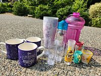Front angled view of entire lot showing mugs, water bottles, tumbler, plastic shooters, assorted shot glasses