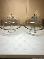 Two clear glass cake stands with domes side by side