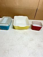 Three colored Pyrex refrigerator dishes with two lids displayed on table