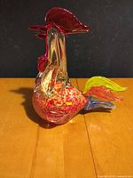 Right side view of blown glass rooster showing red mottled body, clear glass neck, red comb, yellow-green and blue tail