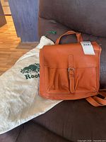 Front view of leather backpack with buckle flap beside Roots dust bag