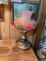 Full view of lamp showing brass-tone base, coiled cord, puffy pink glass shade with roses, clear chimney